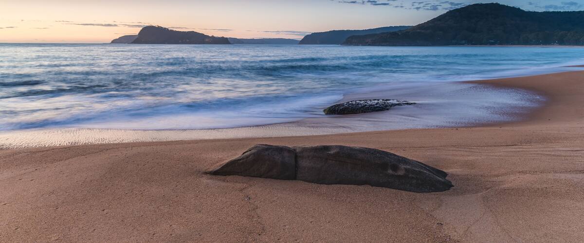 A beach with a large rock in the foreground, calm waves, and distant hills under a twilight sky.