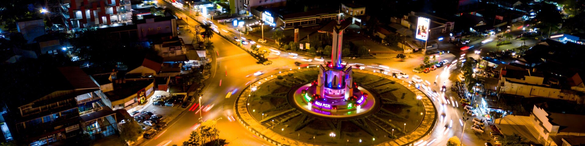 A nighttime aerial view of a city with a central roundabout, illuminated buildings, and a prominent tower.