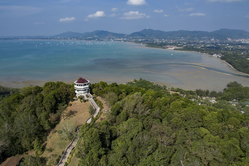 Un paisaje costero con un faro en una colina, una bahía con barcos y una zona boscosa.