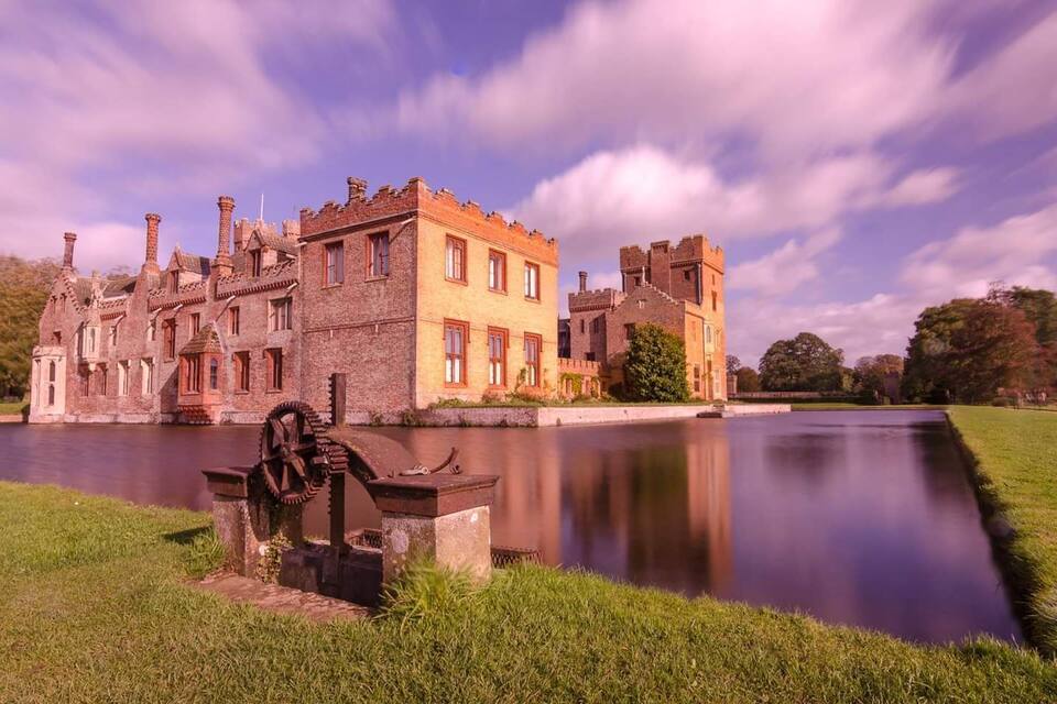 A historic castle with a moat, a large water wheel, and a well-maintained lawn.