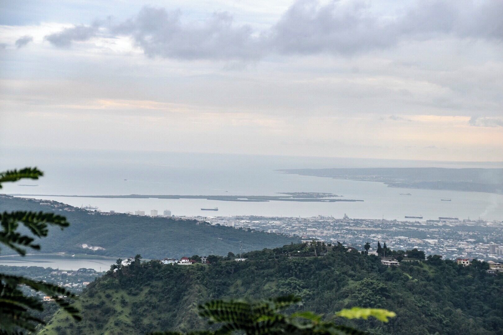 A coastal cityscape with a view of the sea, a distant island, and a hilly terrain with scattered buildings.