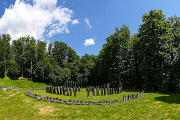 A cemetery with headstones and a circular stone wall, surrounded by lush green trees and a clear blue sky.