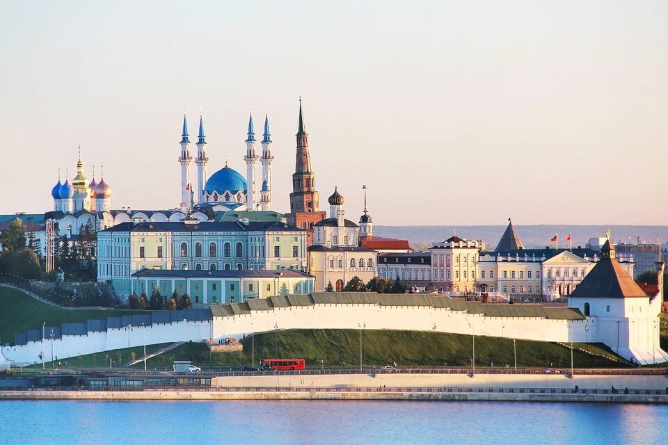 Un paisaje urbano histórico con una catedral destacada, con cúpulas azules y múltiples agujas, un muro blanco con un portón y un río en primer plano.