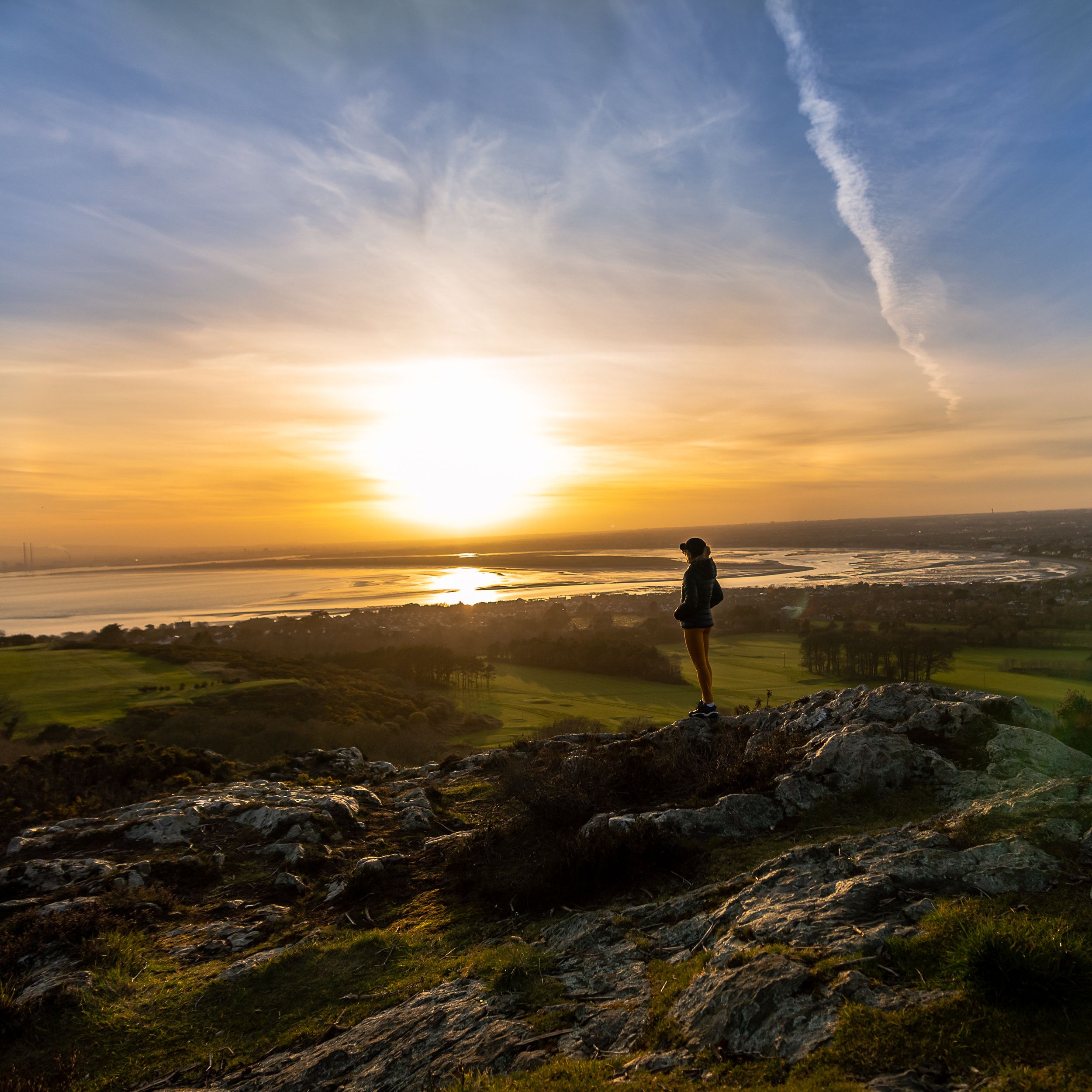 A person standing on a rocky outcrop overlooking a sunset over a body of water with a distant city skyline.
