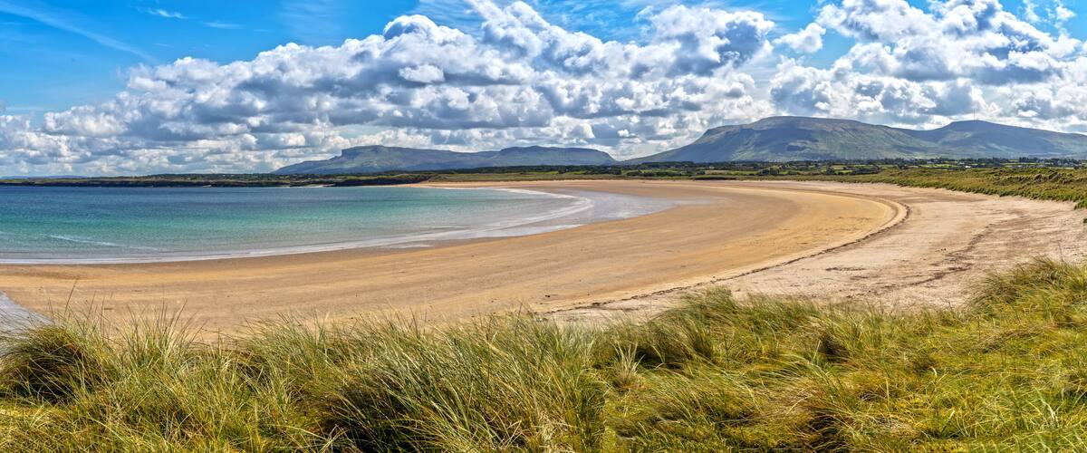 A curved sandy beach with grassy dunes, a calm sea, and distant mountains under a partly cloudy sky.