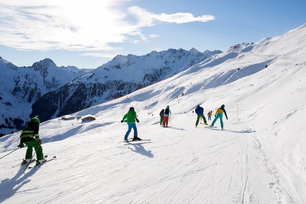 Skiers descending a snowy mountain slope with a clear sky and snow-covered peaks in the background.