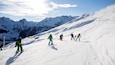 Skiers descending a snowy mountain slope with a clear sky and snow-covered peaks in the background.
