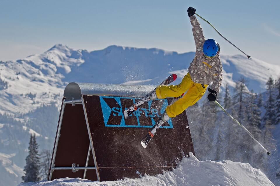 Un skieur en plein saut, réalisant une figure acrobatique sur une rampe enneigée, avec une montagne en arrière-plan.