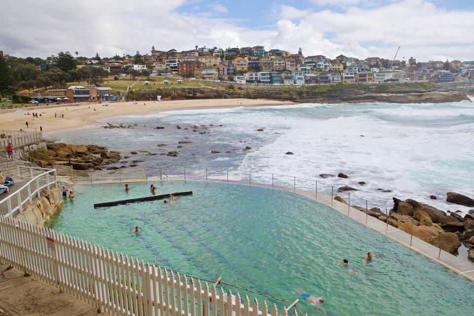 A coastal swimming pool with clear water, surrounded by rocks and a white fence. In the background, there is a beach with people and a hillside of houses.