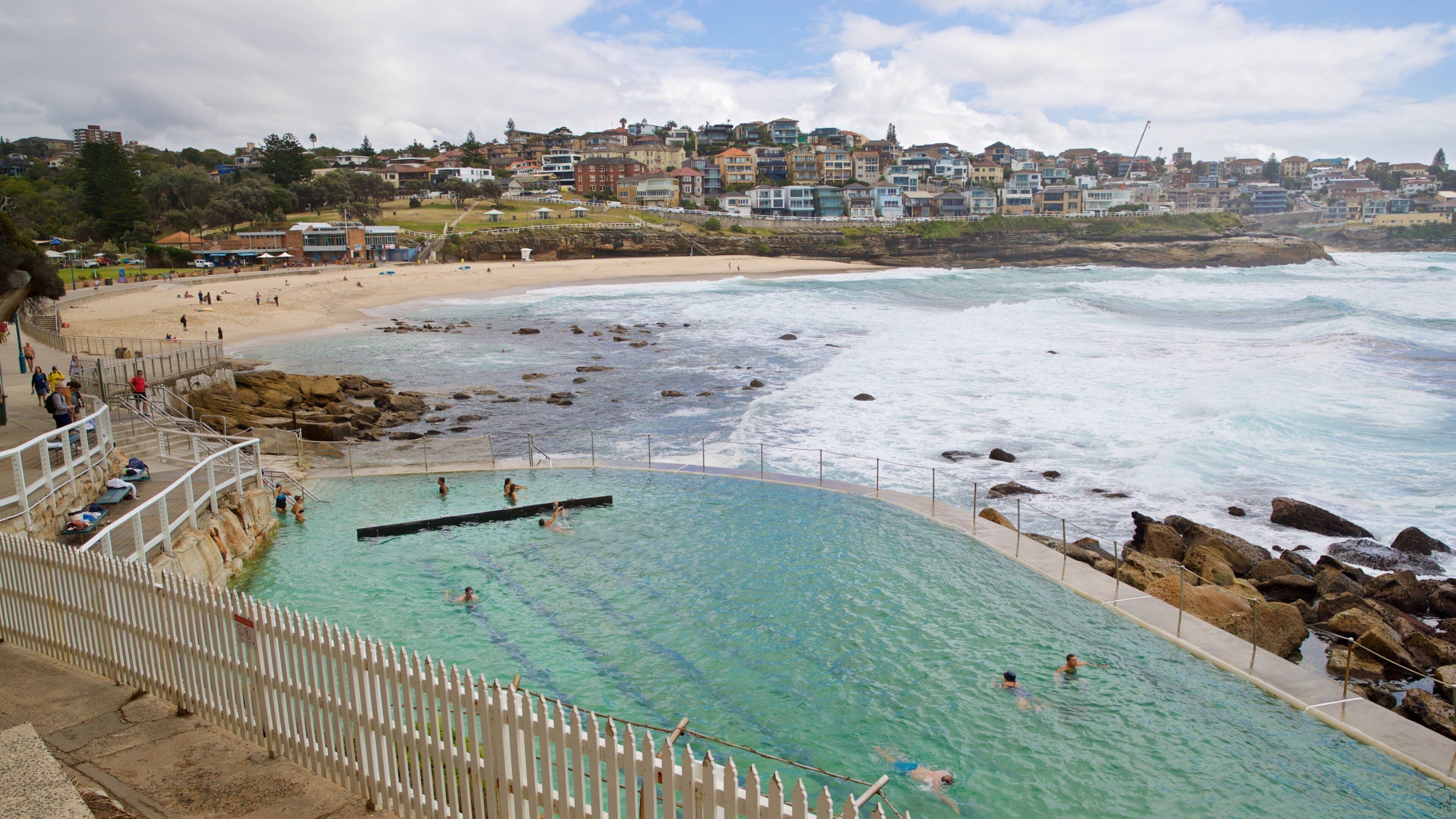 A coastal swimming pool with clear water, surrounded by rocks and a white fence. In the background, there is a beach with people and a hillside of houses.