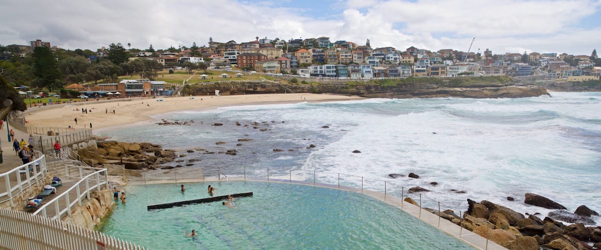 A coastal swimming pool with clear water, surrounded by rocks and a white fence. In the background, there is a beach with people and a hillside of houses.