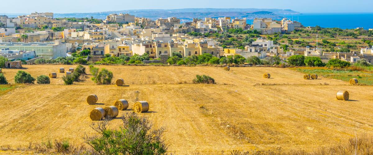 A coastal town with a clear view of the sea, a dry field with hay bales, and a dense cluster of buildings.
