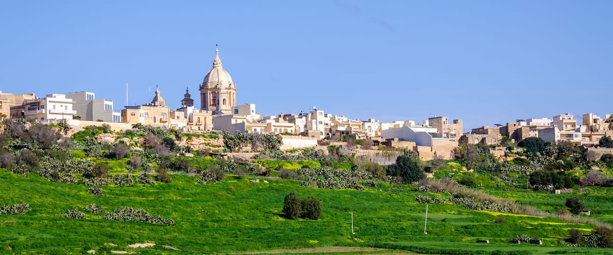 A cityscape with a prominent dome-shaped building, lush green fields, and a clear blue sky.