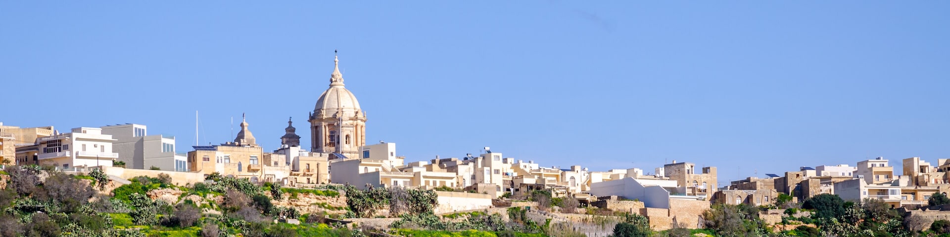 A cityscape with a prominent dome-shaped building, lush green fields, and a clear blue sky.