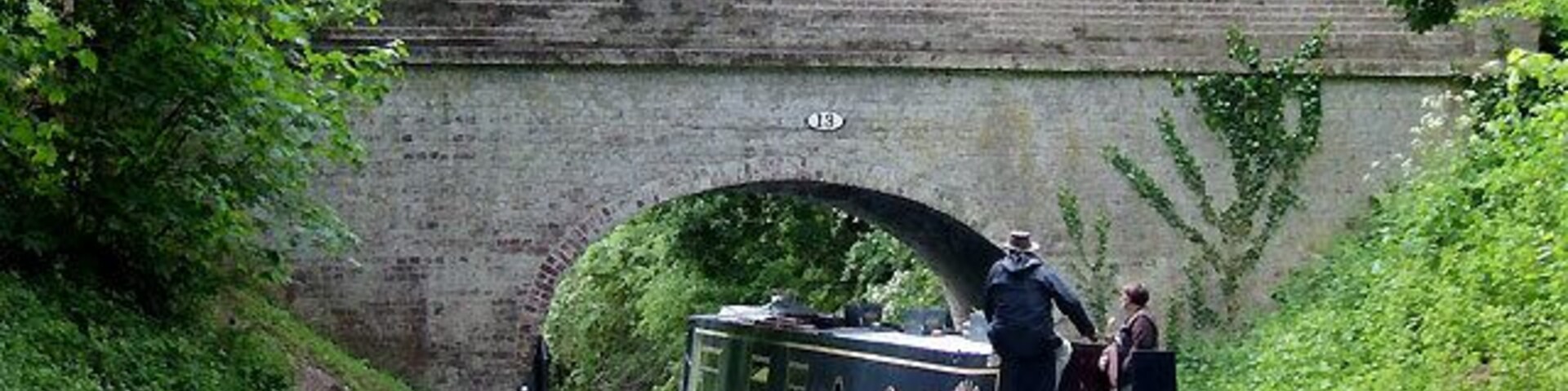 Shropshire Union Canal Bridge No 13 at Brewood, Staffordshire. Narrowboat "Swallow" negotiates School Bridge at Brewood. The canal was built by Thomas Telford and opened in 1835. He attempted to minimise the distance by adopting straight lines wherever possible, so his canal includes many long embankments, and cuttings such as the one here at Brewood.