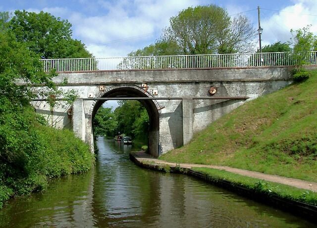 A stone bridge over a narrow canal with a pathway on the right and trees in the background.
