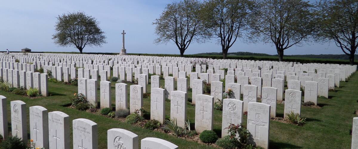 A cemetery with numerous headstones, a central cross monument, and trees in the background.