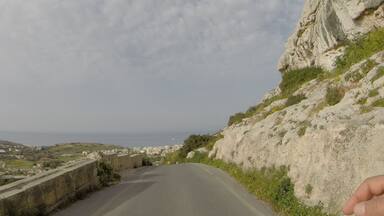 A winding road with a rocky hillside on the right and a coastal town in the distance.