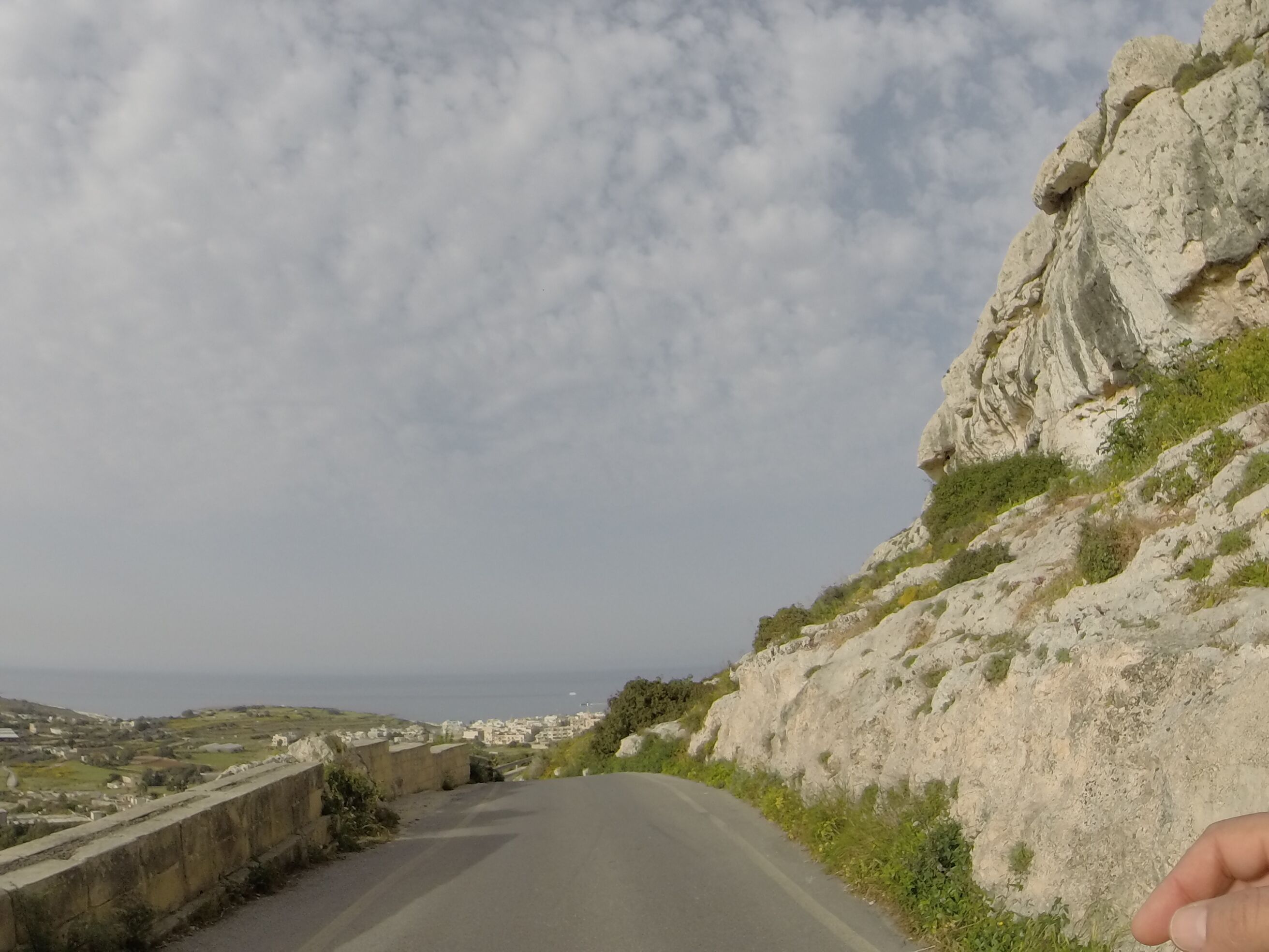 A winding road with a rocky hillside on the right and a coastal town in the distance.