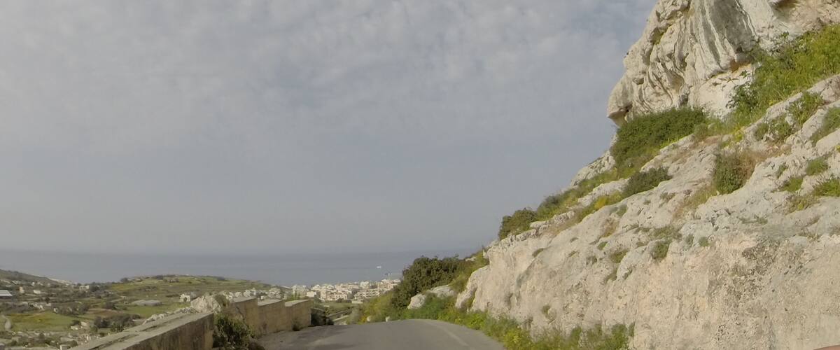A winding road with a rocky hillside on the right and a coastal town in the distance.