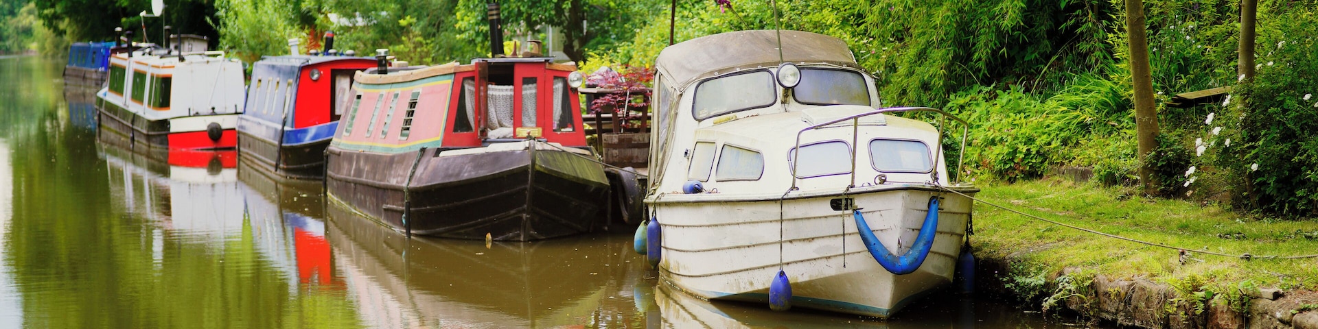 The picture shows a colourful narrow boat / barge / houseboat moored on the Grand Union Canal in Warwickshire, in the West Midlands, England, UK. It is an afternoon in summer and there is hazy sunshin
