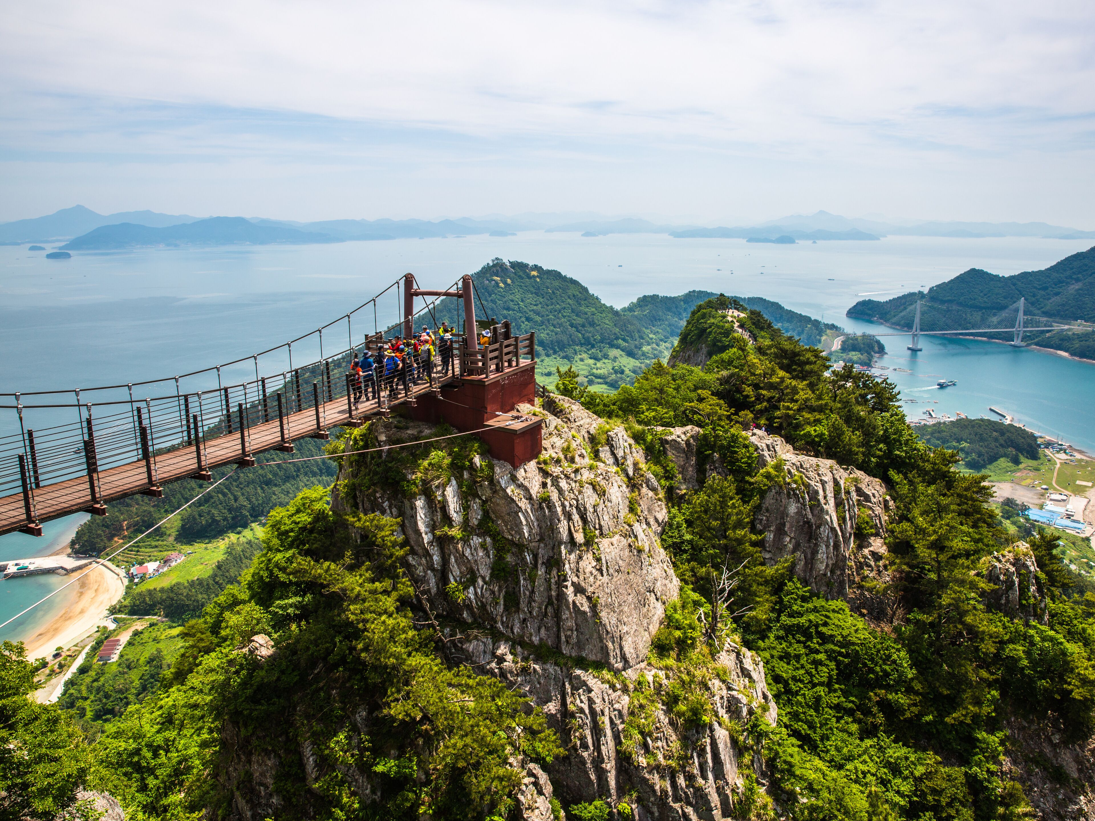 Un puente colgante con gente caminando, con vistas a una zona costera con montañas y un cielo despejado.