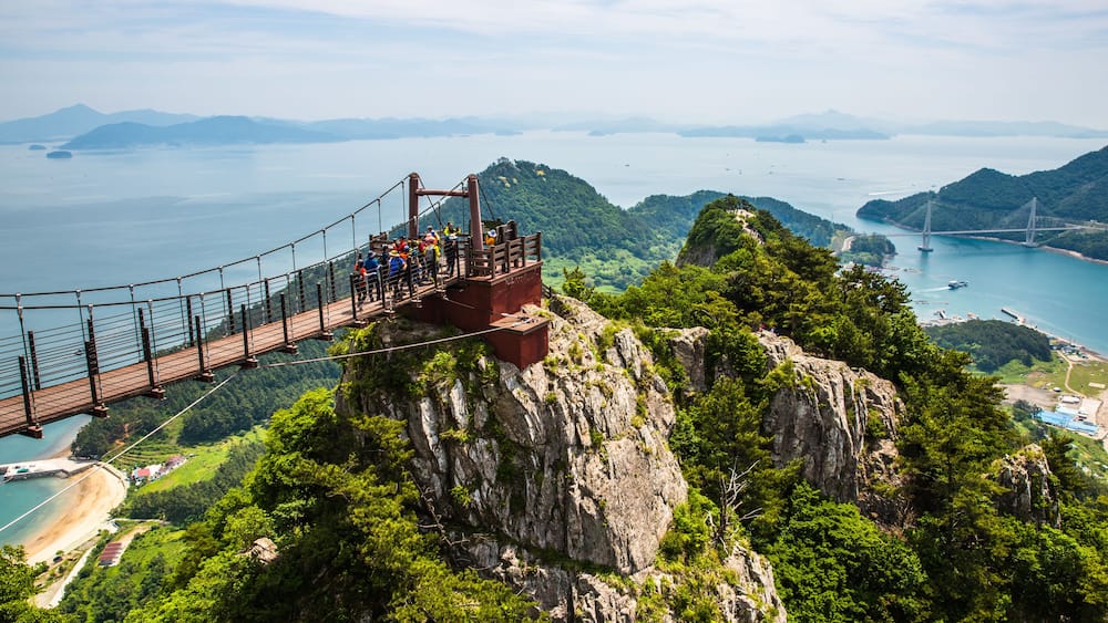 A suspension bridge with people walking across it, overlooking a coastal area with mountains and a clear sky.