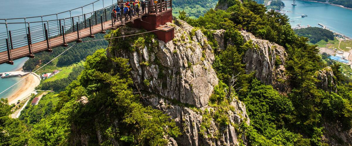 Un puente colgante con gente caminando, con vistas a una zona costera con montañas y un cielo despejado.