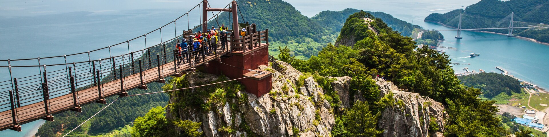 Un puente colgante con gente caminando, con vistas a una zona costera con montañas y un cielo despejado.