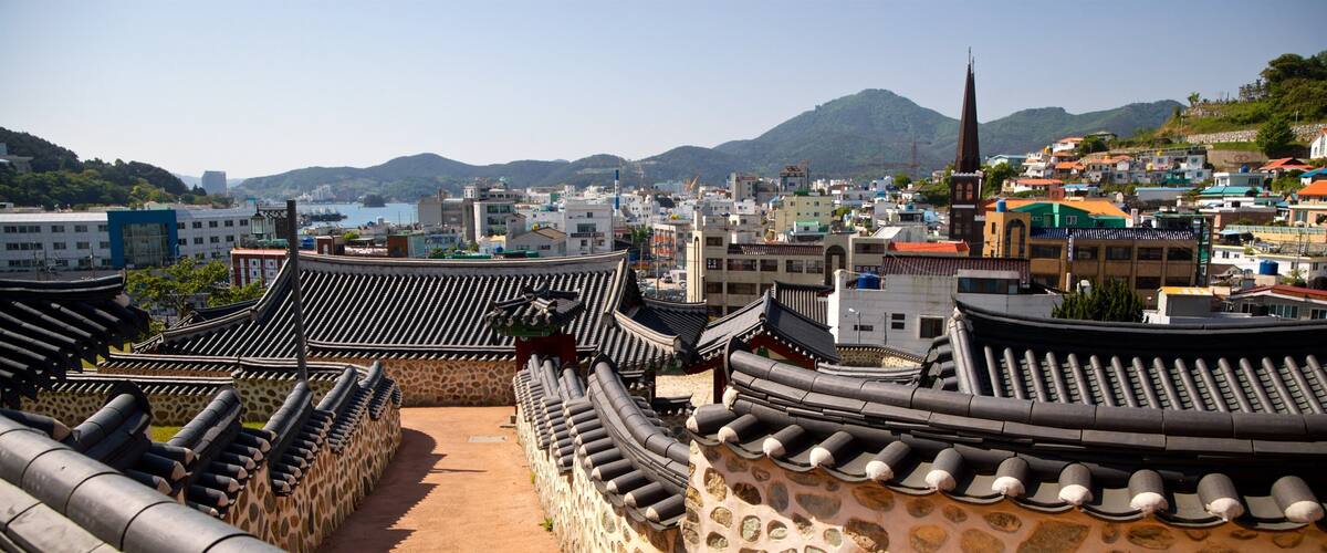 Traditional Korean architecture with curved roofs and stone walls, overlooking a coastal cityscape with mountains in the background.