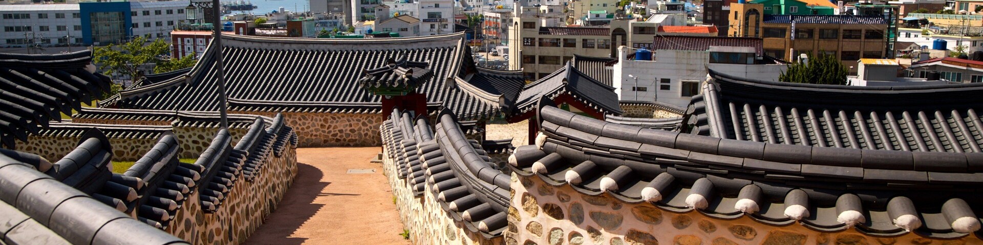 Traditional Korean architecture with curved roofs and stone walls, overlooking a coastal cityscape with mountains in the background.
