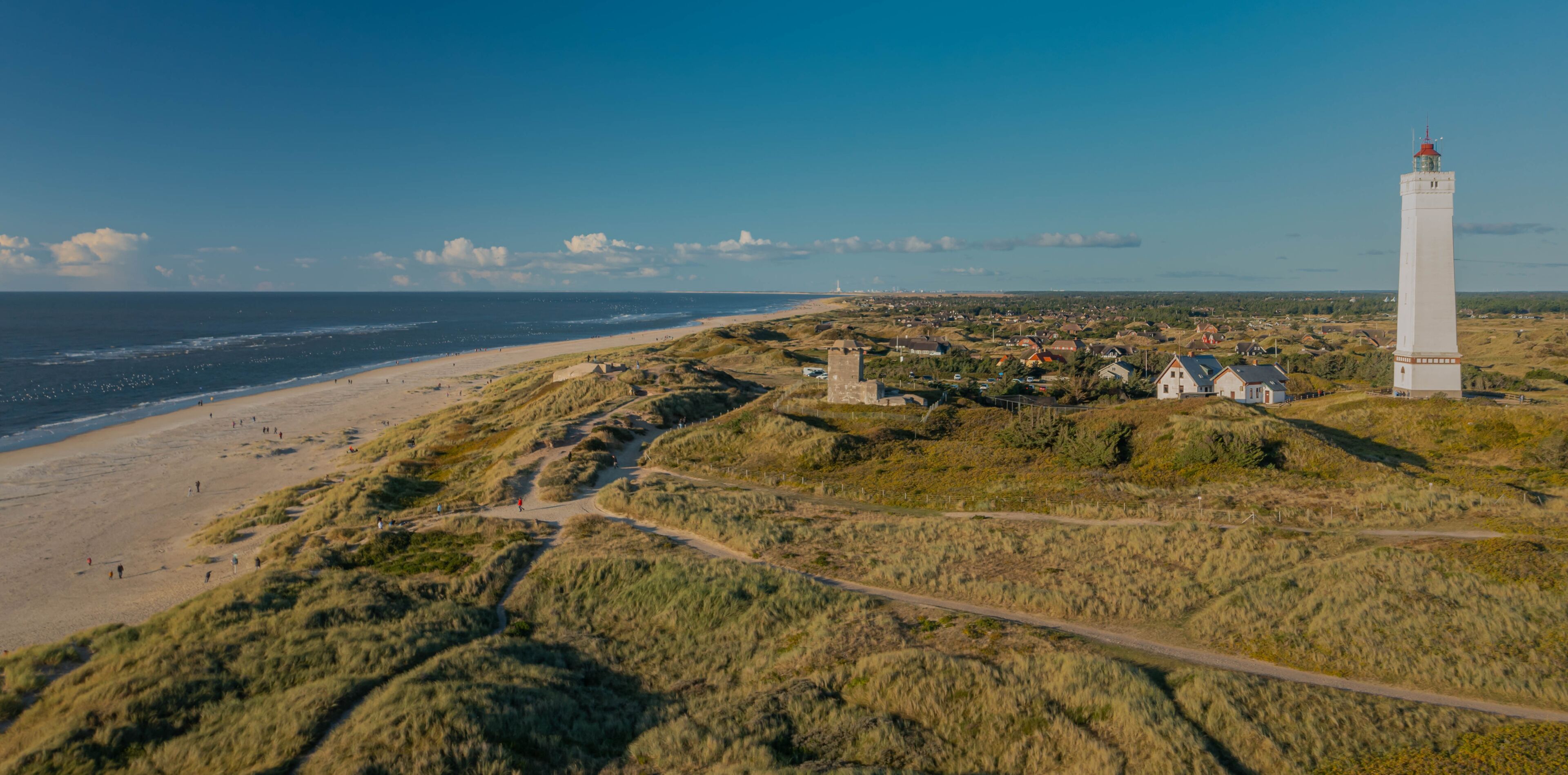 Un paisaje costero con un faro, playa, dunas y un pueblo pequeño.