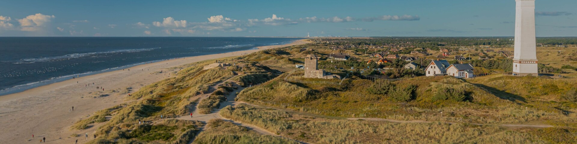 Un paisaje costero con un faro, playa, dunas y un pueblo pequeño.