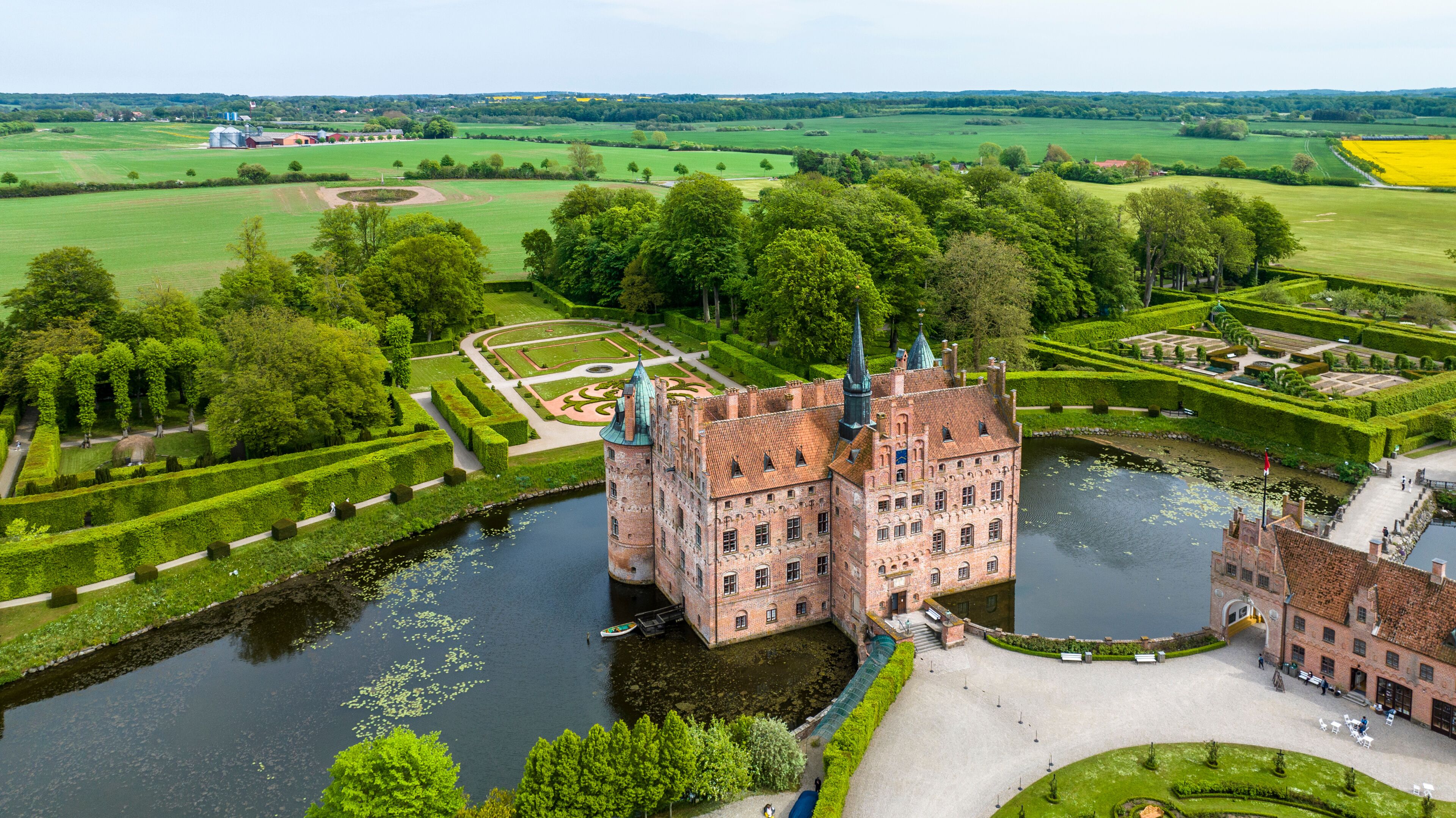 Um castelo histórico com um fosso, rodeado por jardins bem cuidados e campos.