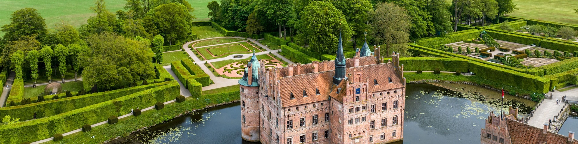 Um castelo histórico com um fosso, rodeado por jardins bem cuidados e campos.