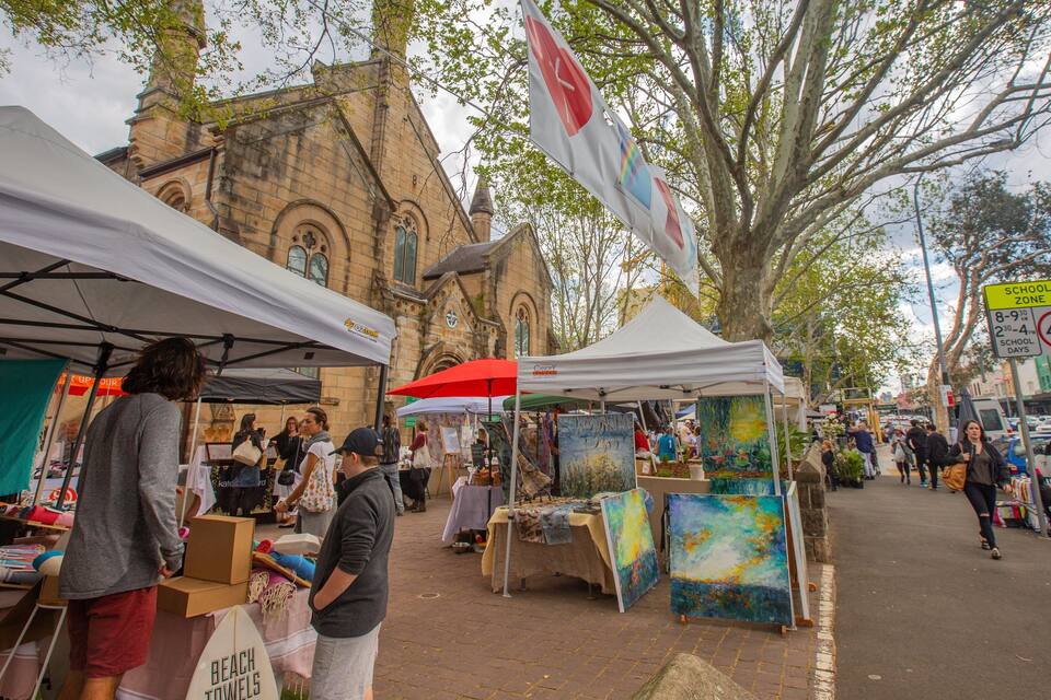 Un mercado al aire libre con puestos que venden toallas de playa y arte, frente a un edificio histórico y una calle arbolada.