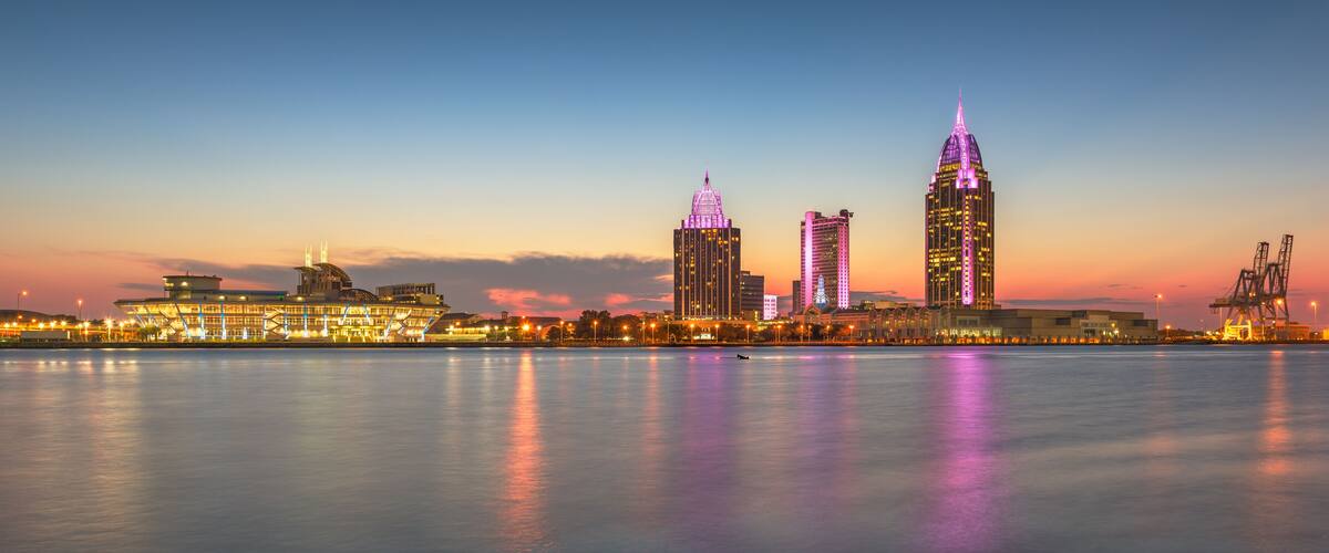 A city skyline at dusk with illuminated buildings reflecting on the water.
