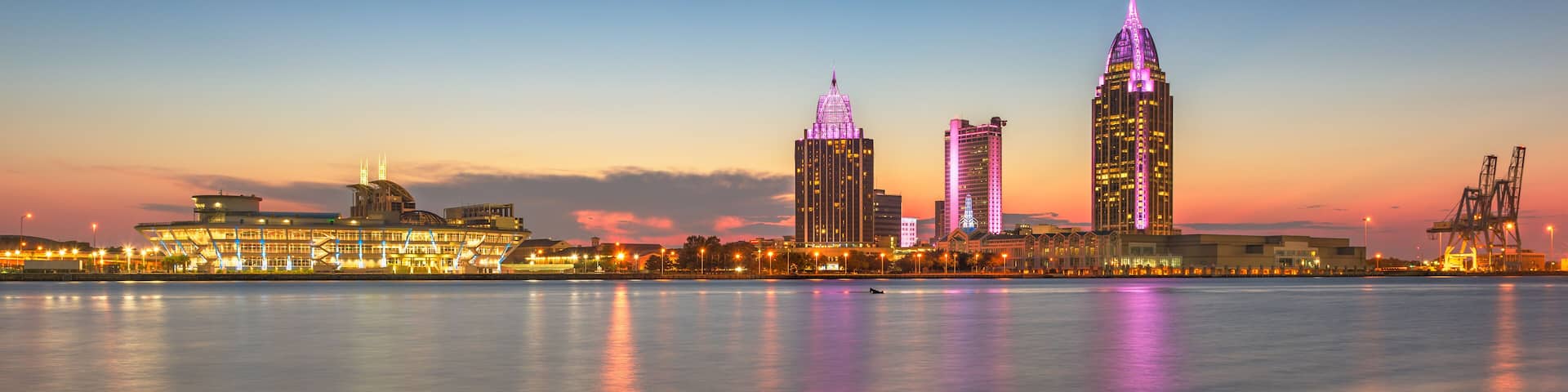 A city skyline at dusk with illuminated buildings reflecting on the water.