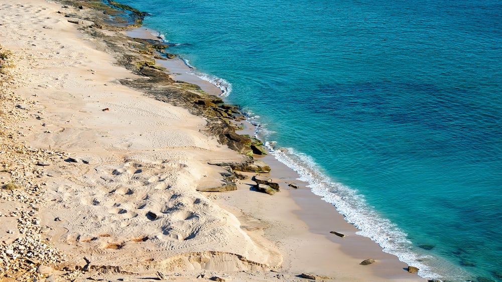 A sandy beach with clear blue water and rocky outcrops.