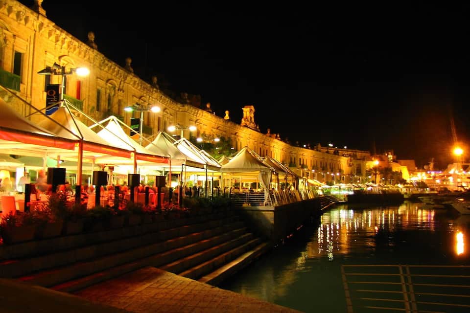 Une promenade en bord de mer, illuminée la nuit, avec des bâtiments éclairés et un bateau amarré au bord de l’eau.
