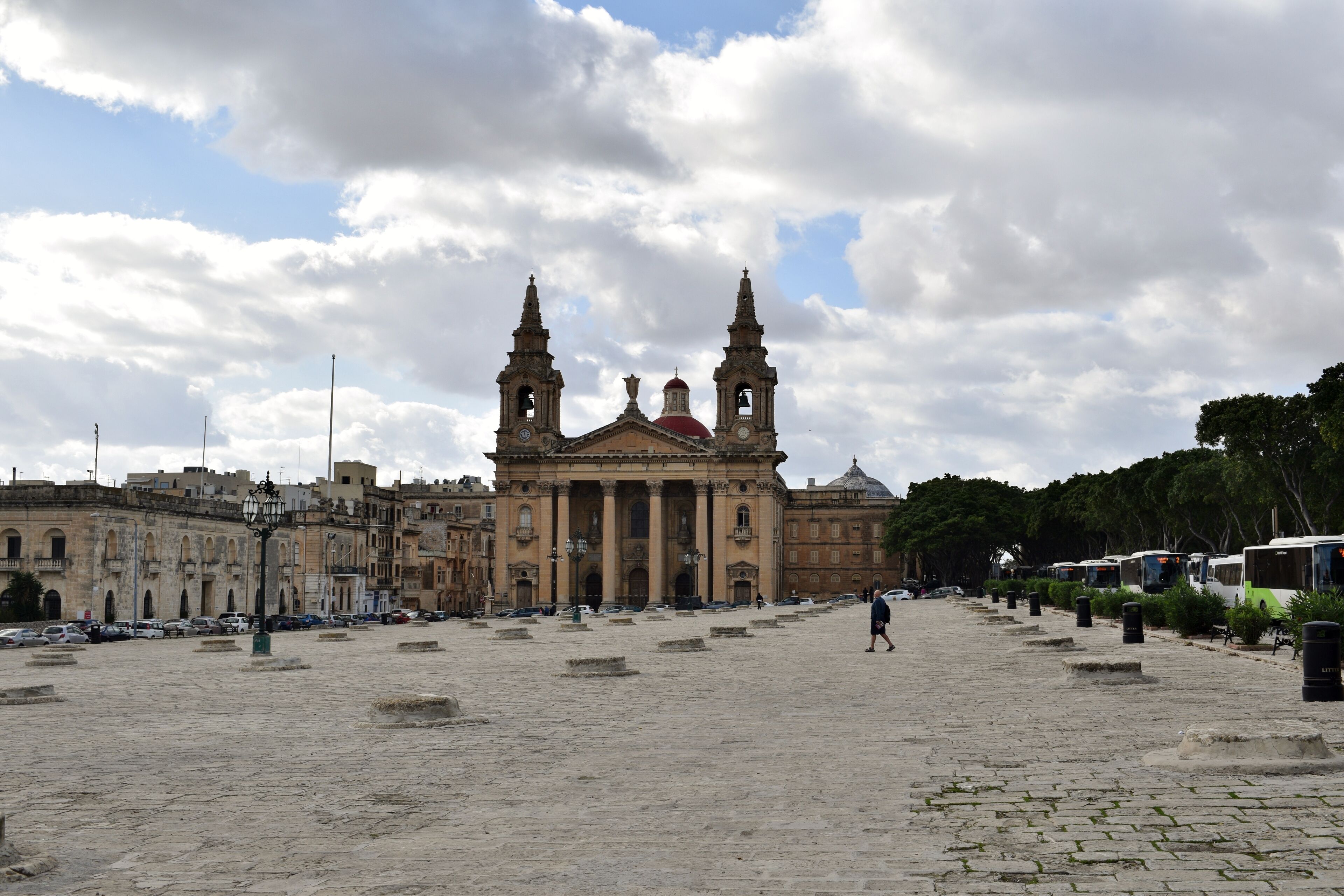 A historic building with two towers and a red dome, surrounded by a cobblestone plaza with parked buses.
