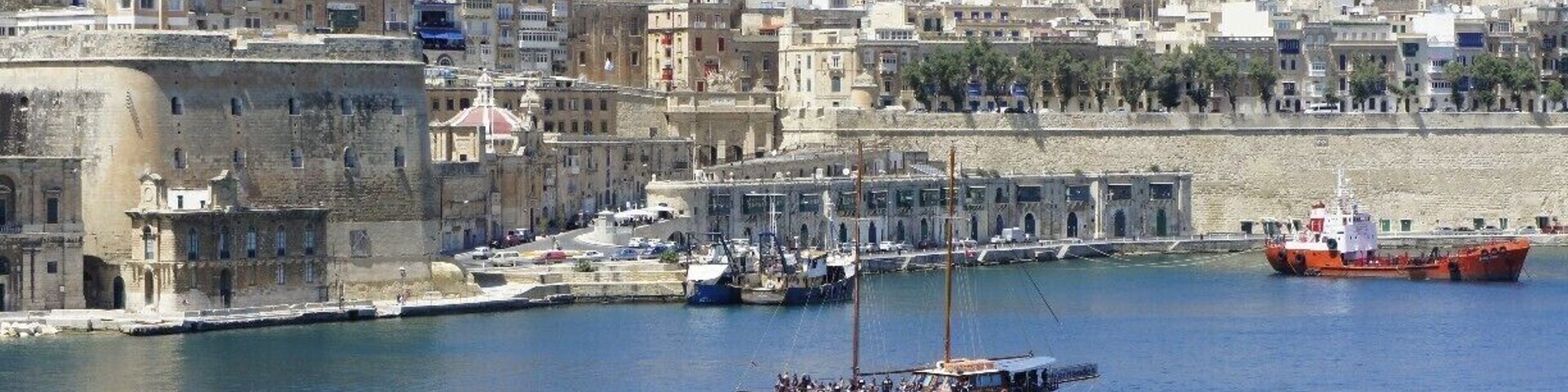 A coastal cityscape with historic buildings, a harbor with boats, and a clear blue sky.