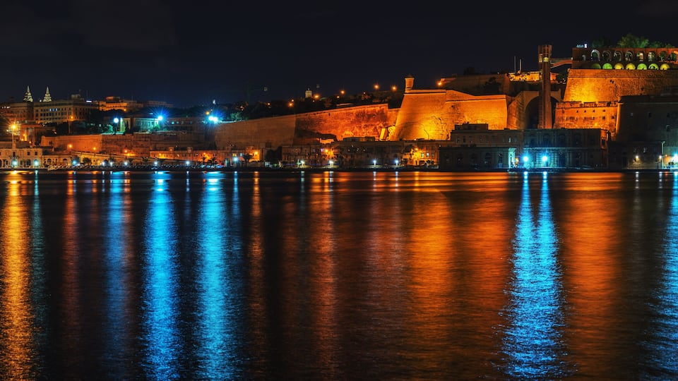 A nighttime cityscape with illuminated buildings reflecting on the water.