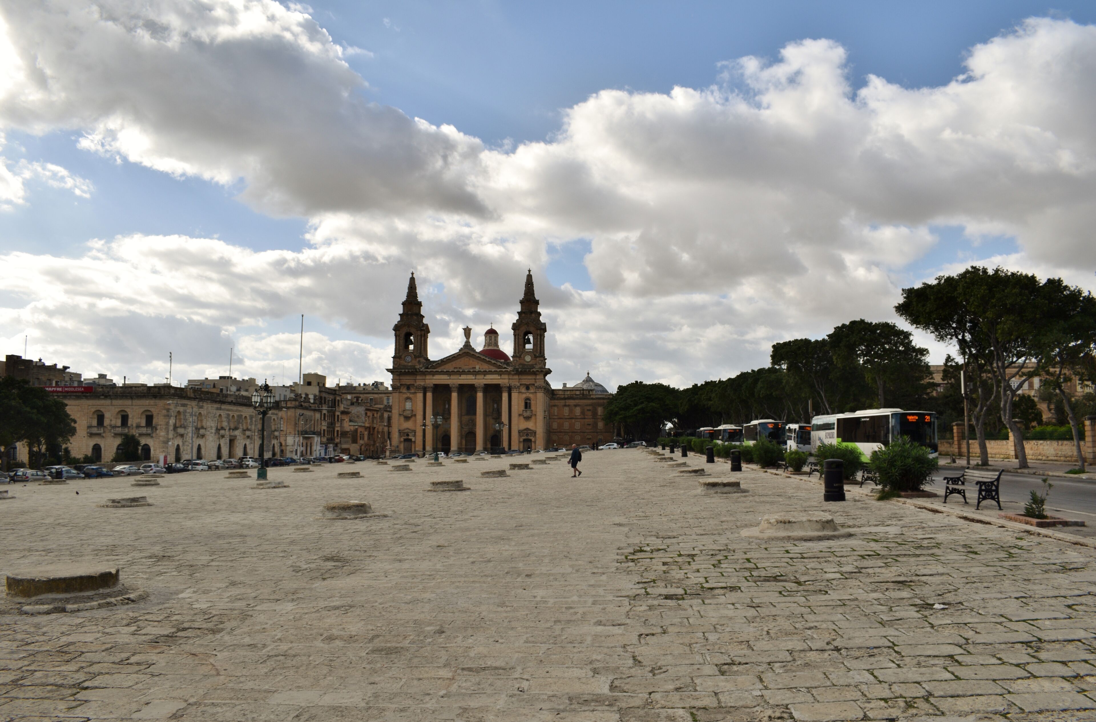 A historic plaza with a grand building featuring two towers, surrounded by trees and a paved walkway.