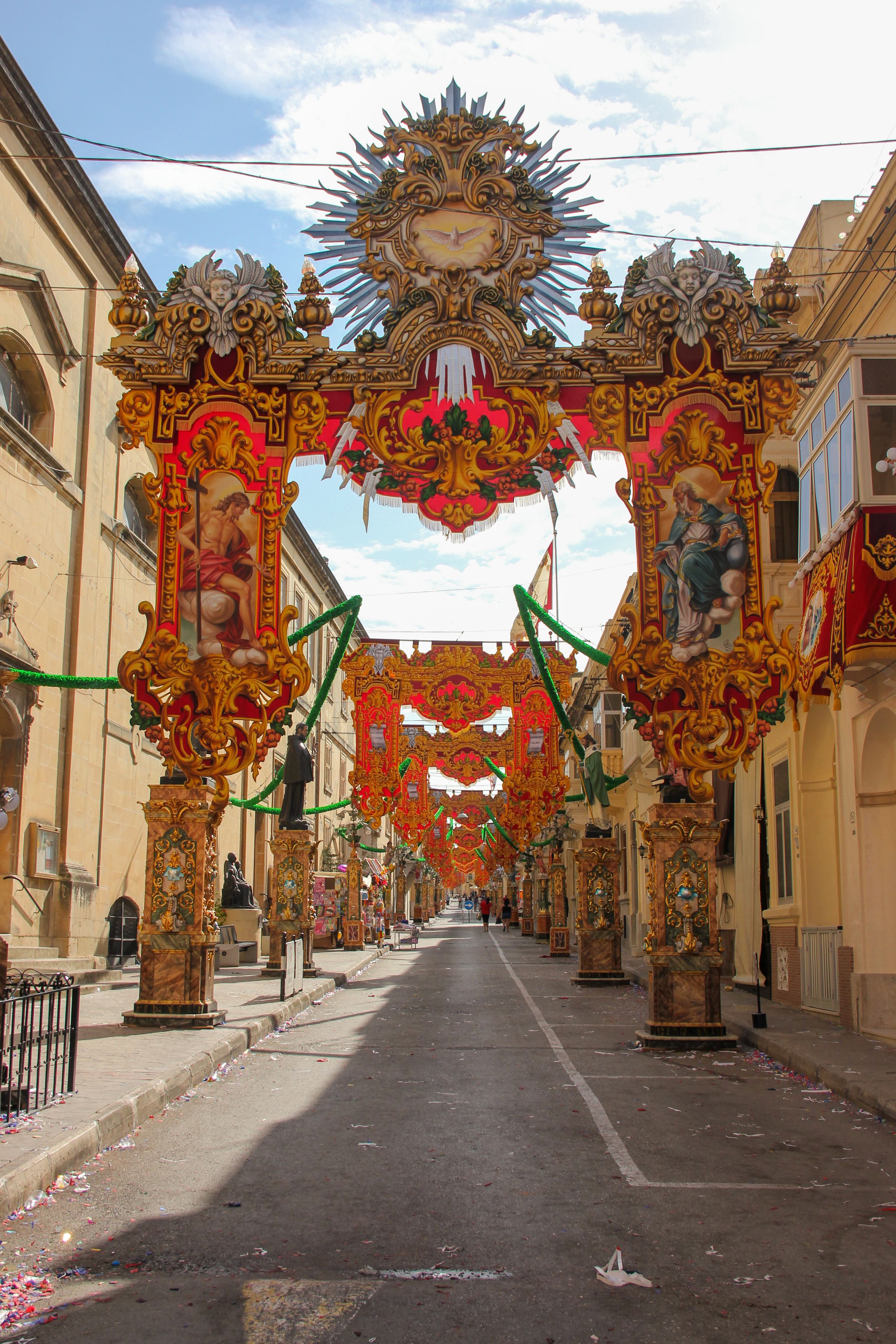 A street adorned with ornate golden arches and statues, leading to a building with a red facade.