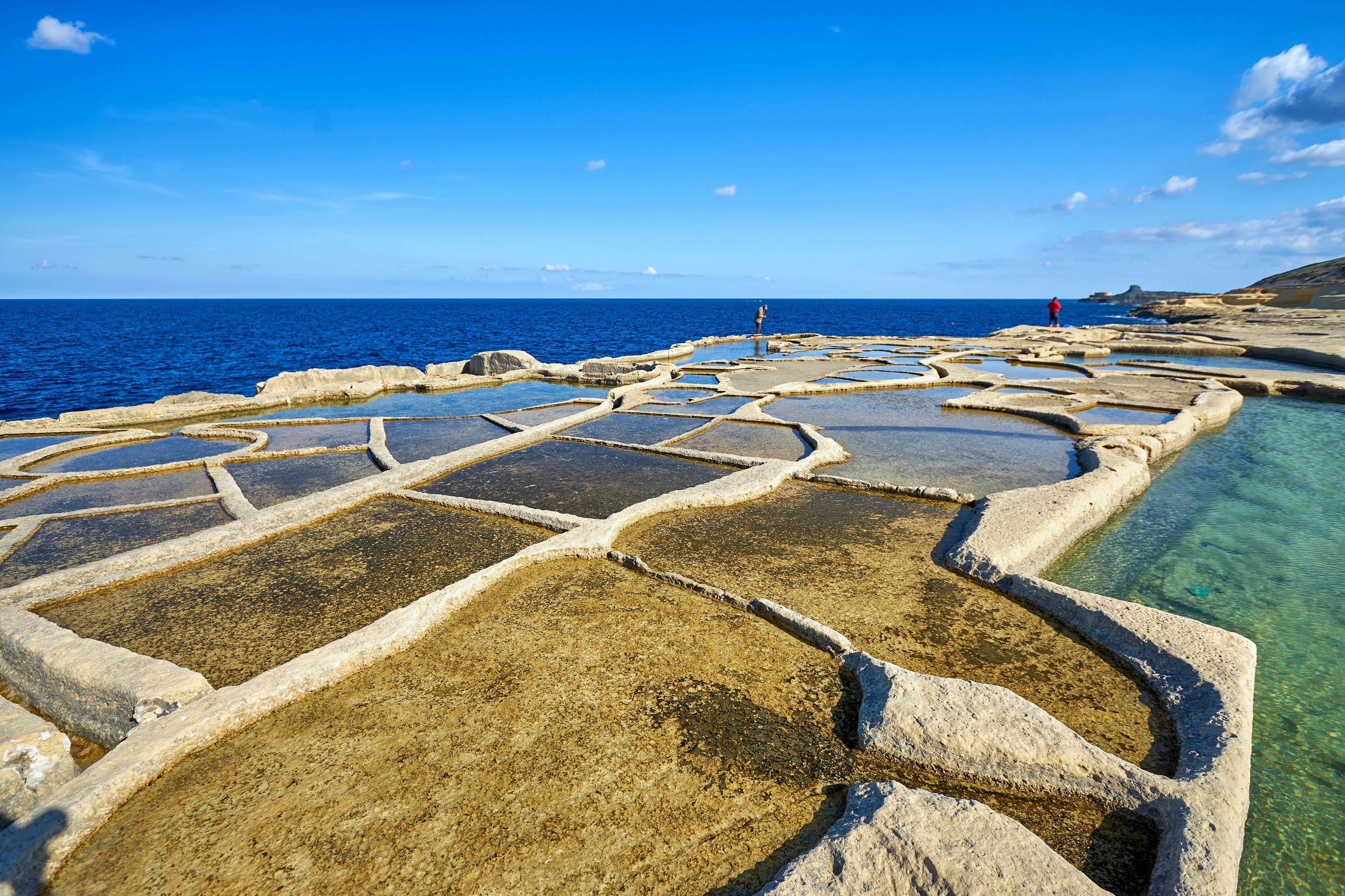 Paesaggio costiero con piscine a terrazze di acqua limpida, una persona in piedi a destra e l'oceano sullo sfondo.
