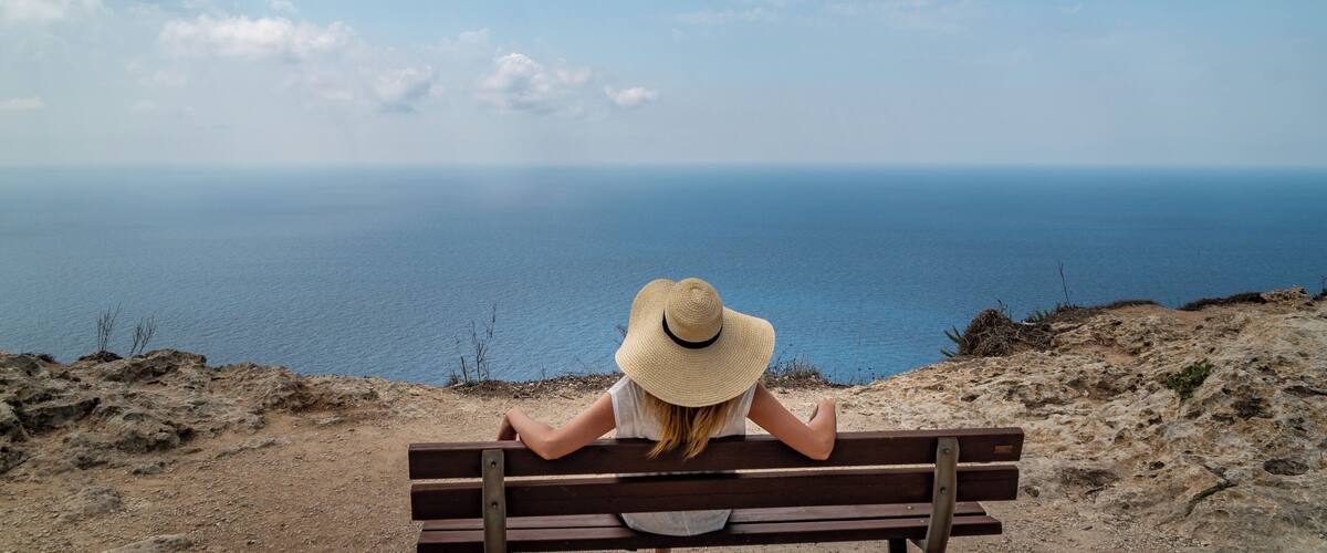 A woman in a straw hat sitting on a bench overlooking a coastal view.