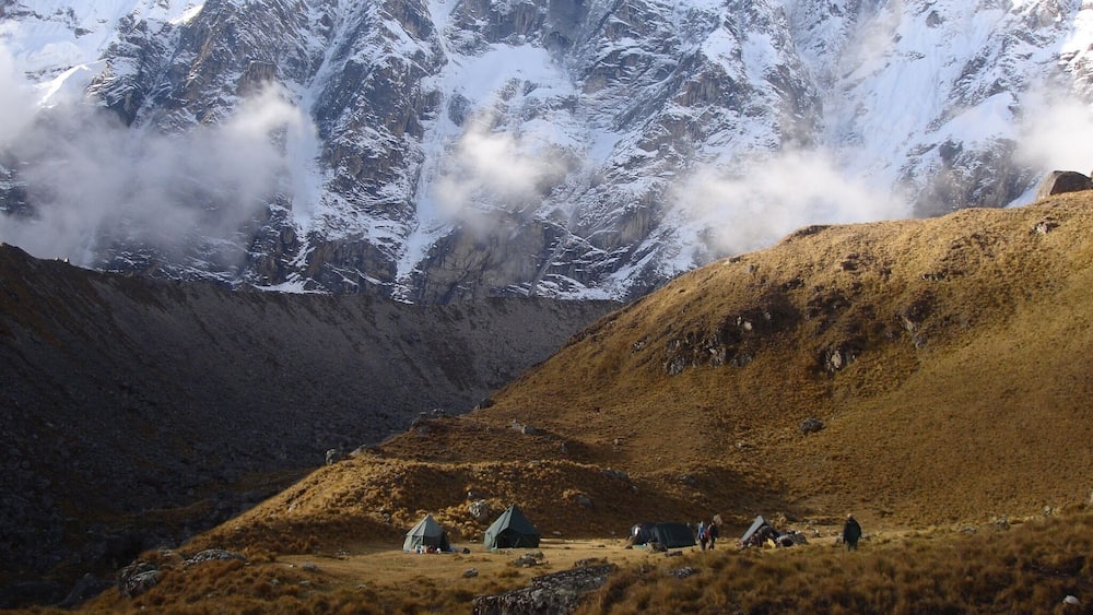 Un paysage montagneux aux sommets enneigés, une vallée parsemée de tentes et une personne qui marche.