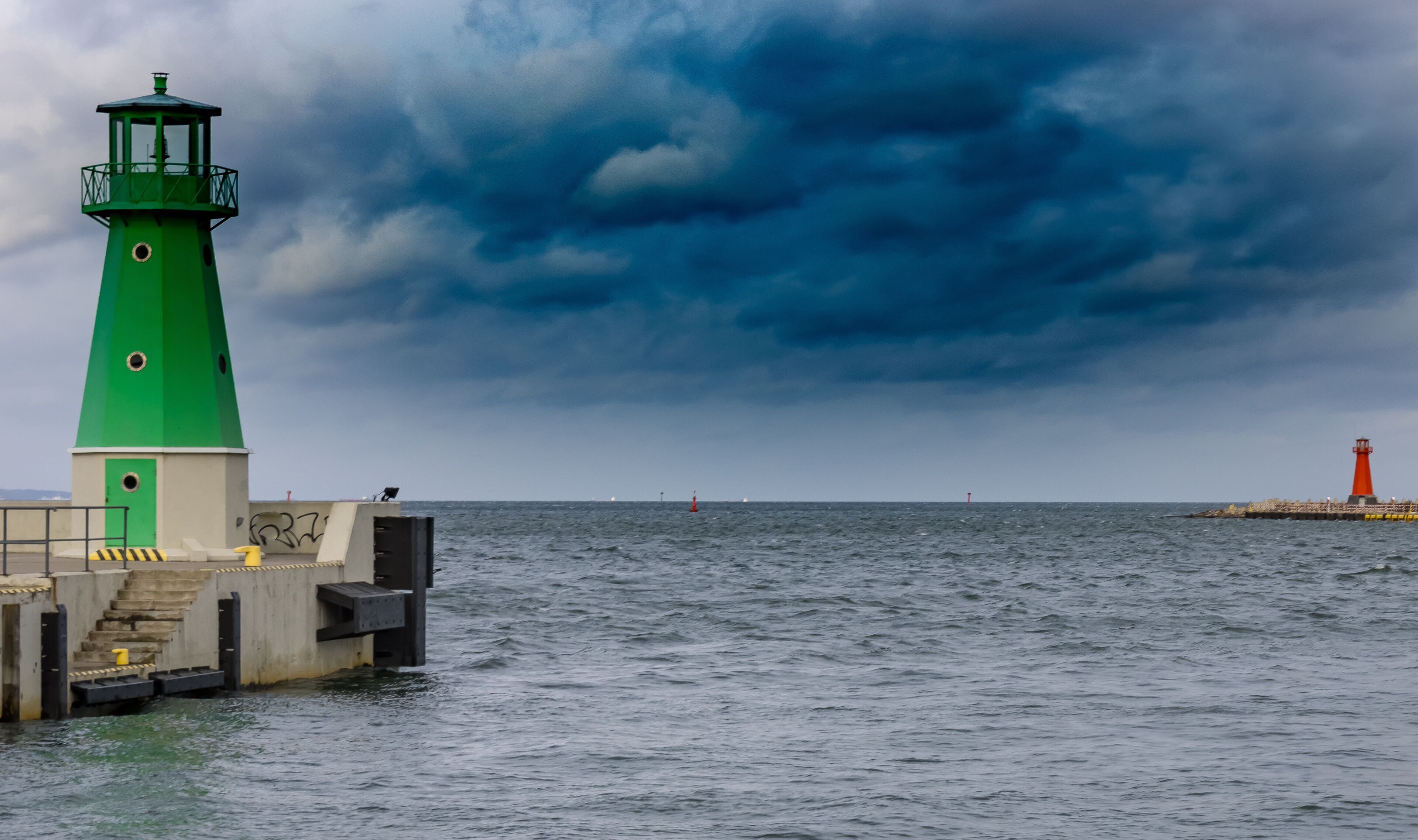 A green lighthouse on a pier with a red lighthouse in the distance.