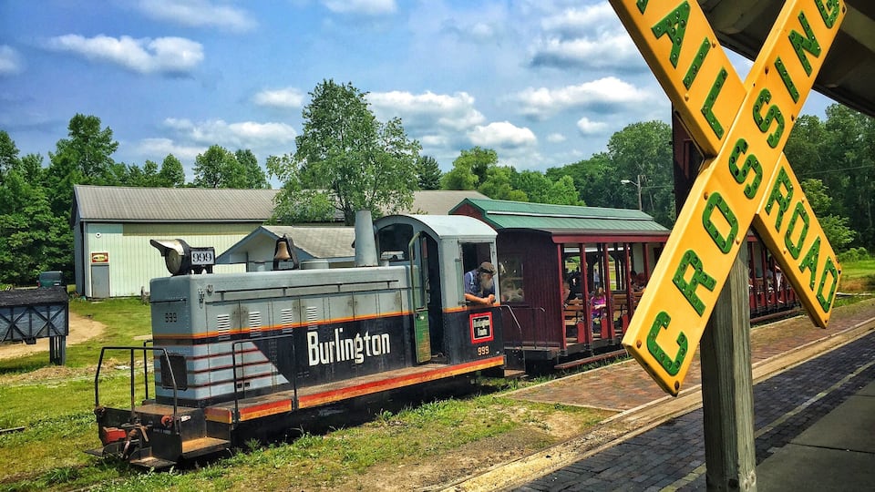 A Burlington train at a railway crossing with a vintage train car and a station building in the background.